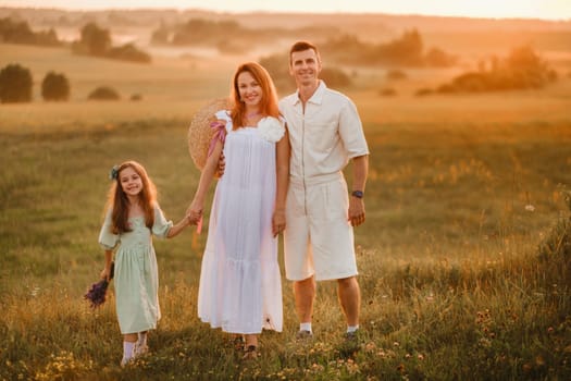 A beautiful happy family of three is standing in a field at sunset.