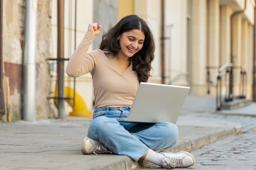 Overjoyed happy young Indian woman working on laptop scream in delight raise hands in triumph winner gesture celebrate success game money win outdoors. Happy girl sitting on downtown street in city