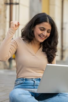 Overjoyed happy Indian woman working on laptop scream in delight raise hands in triumph winner gesture celebrate success game money win outdoor. Happy girl sitting on downtown street in city. Vertical