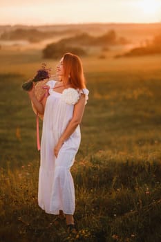 A girl in a white dress with a bouquet of lavender flowers stands in a field at sunset.