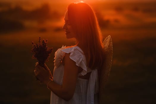 A girl in a white dress with a bouquet of lavender flowers stands in a field at sunset.