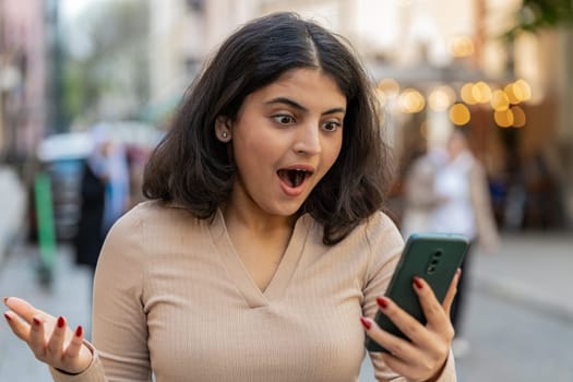 Oh my God Wow. Excited happy joyful winner Indian young woman use smartphone typing browsing celebrating win good message news outdoors. Hispanic girl walking on urban city street. Town lifestyles