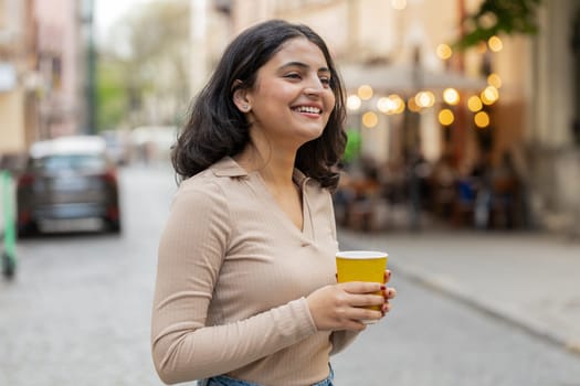 Smiling happy Indian young woman enjoying morning coffee hot drink outdoors. Relaxing, taking a break. Hispanic girl drinking coffee to go walking passes the urban city street. Town lifestyles outside