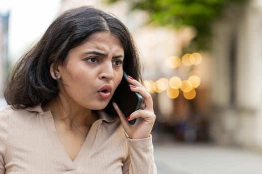 Angry mad stressed Indian young woman arguing while talking on smartphone irritated shout quarrel outdoors. Hispanic offended dissatisfied angry girl standing on urban city street. Town lifestyles