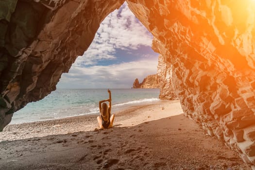 Woman travel sea. View of a woman in a black swimsuit from a sea cave Attractive woman enjoying the sea air sits on the beach and looks at the sea. Behind her are rocks and the sea.