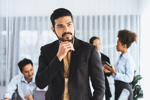 Businessman poses confidently with diverse coworkers in busy meeting room background. Multicultural team works together for business success. Modern businessman portrait. Concord