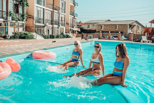 Three women are sitting in a pool with pink and blue floaties. Scene is lighthearted and fun