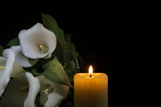 Close-up view of a single burning wax candle next to white calla lily flower with a green stem and leaves, contemplative and serene atmosphere, suggesting themes of remembrance, reflection and hope