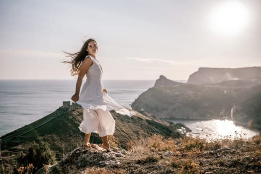 A woman stands on a hill overlooking the ocean. She is wearing a white dress and has long hair. The scene is serene and peaceful, with the sun shining brightly in the background