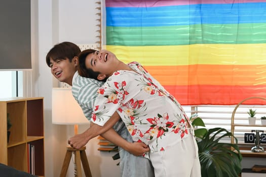 Playful gay couple giving piggyback ride for each other laugh joyfully in living room near rainbow LGBT Pride flag.