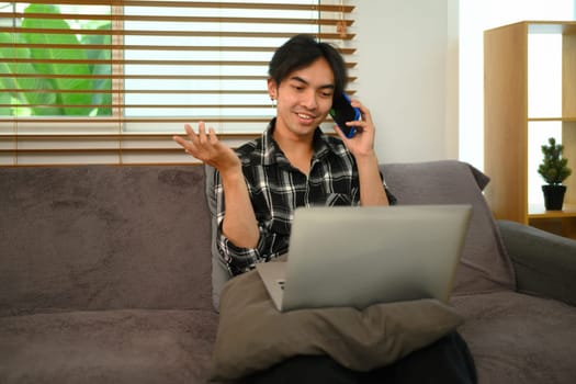 Smiling male freelancer having pleased phone conversation and using laptop on couch at home.