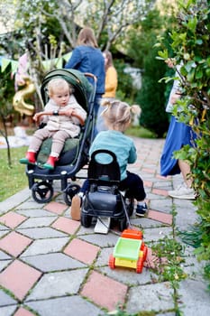 Small child sits in a stroller and looks at a little girl driving a toy car with a small truck tied to the seat. High quality photo