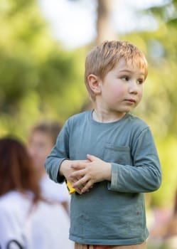 A young boy wearing a green shirt and brown pants is holding a yellow object in his hand. He looks up at the camera with a curious expression