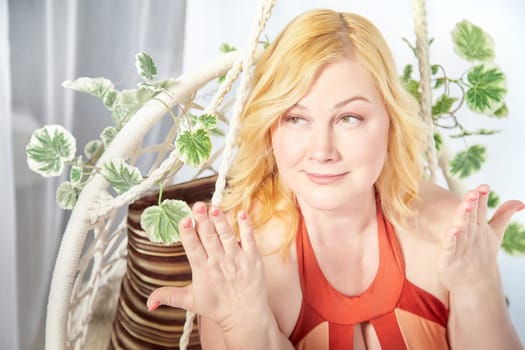 Portrait of Funny cute mature adult woman with blonde hair posing in swimsuit in a wicker chair with plants. Fat middle-aged lady in underwear in studio. The concept of body positivity and happy
