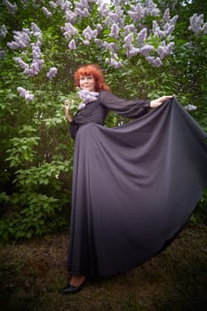 Elegant senior mature Woman in Black Dress looking like a witch by Blooming Lilac Bush at Dusk. Woman with red hair stands poised among lilac blooms