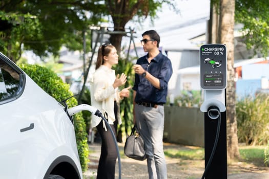 Young couple recharge electric car's battery from charging station in outdoor green city park in springtime. Rechargeable EV car for sustainable environmental friendly urban travel lifestyle.Expedient