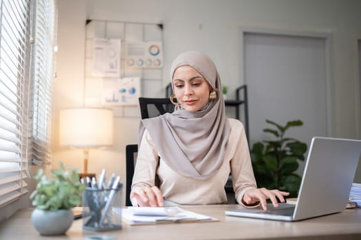 Young Muslim businesswoman wearing hijab sits working with laptop managing personal business in private office..