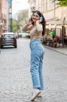 Rear view of happy Indian young woman in wireless headphones dancing listening favorite energetic disco music in smartphone dancing outdoors. Hispanic girl walking on urban city street. Vertical