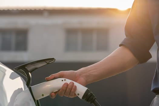 Young man put EV charger to recharge electric car's battery from charging station in city commercial parking lot with sunlight. Rechargeable EV car for sustainable eco friendly urban travel. Expedient