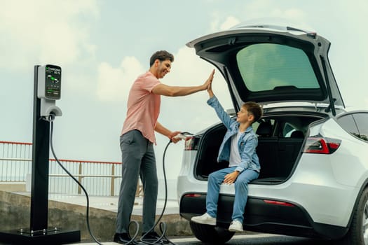 Family road trip vacation traveling by the sea with electric car, father and son high five after reach destination at EV charging station by the seashore. Eco-friendly car for environment. Perpetual