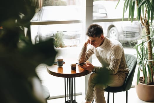 A young man sits at a table in a well-lit, contemporary cafe, deeply focused on his smartphone. He is casually dressed in a sweater and is holding a cup of coffee, enjoying a quiet moment by himself amidst the indoor greenery.
