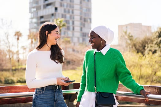 two young female friends smiling happy while chatting carefree in a city park, concept of diversity and modern lifestyle