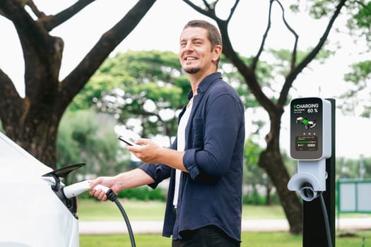 Man using smartphone online banking application to pay for electric car battery charging from EV charging station during vacation holiday road trip at national park or summer forest. Exalt