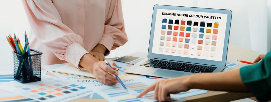 Creative graphic design presents selective color while manager using laptop comparing suitable color at table with color palettes, graphic material scatter around at modern office. Variegated.