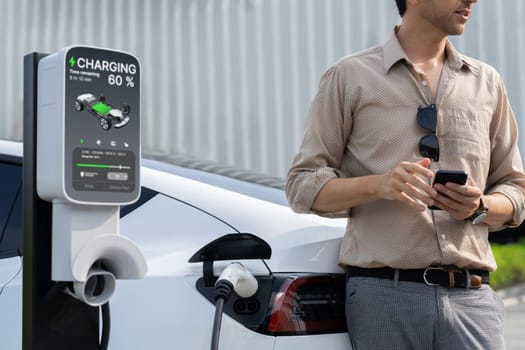 Young man use smartphone to pay for electricity at public EV car charging station at city commercial mall parking lot. Modern environmental and sustainable urban lifestyle with EV vehicle. Expedient