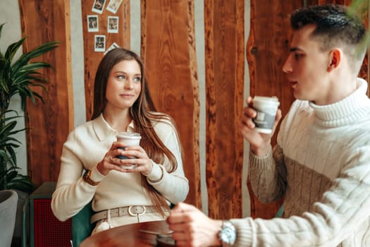 A young man and woman are seated opposite each other in a warm, wood-paneled cafe, each holding a cup of coffee. The woman listens attentively, her gaze fixed on her companion, while the man gestures mid-conversation, creating a relaxed and intimate atmosphere.
