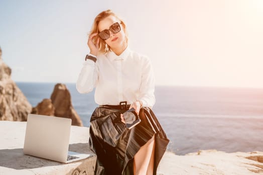 Successful business woman in yellow hat working on laptop by the sea. Pretty lady typing on computer at summer day outdoors. Freelance, travel and holidays concept.