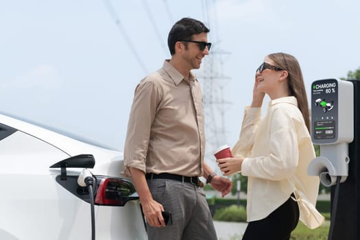 Young couple with coffee recharge EV car battery at charging station connected to electrical industrial power grid tower. Couple with shopping and travel using eco electric car lifestyle. Expedient