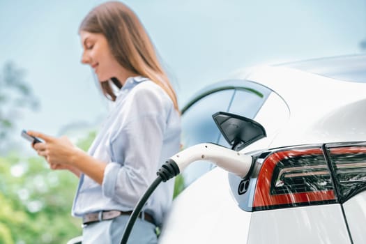 Young woman using smartphone online banking application to pay for electric car battery charging from EV charging station during vacation holiday road trip at national park or summer forest. Exalt