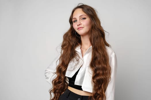 A young woman with flowing, wavy brown hair showcases her casual fashion sense, wearing an unbuttoned white shirt over a black crop top, paired with a black bottom. Her pose exudes confidence as she gazes off-camera, standing elegantly against a plain gray background.