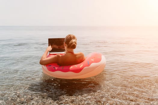 Woman freelancer works on laptop swimming in sea on pink inflatable ring. Pretty lady typing on computer while floating in the sea on inflatable donut at sunset. Freelance, remote work on vacation