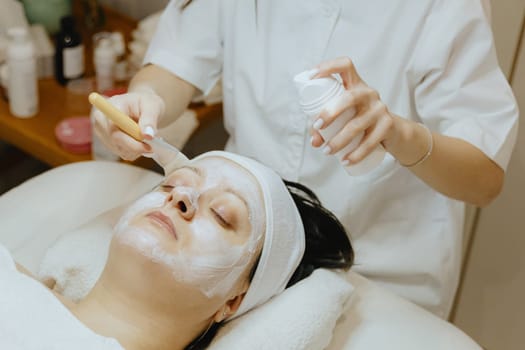 A young beautiful Caucasian brunette girl, a cosmetologist, smears a white cream mask with a brush on the face of an adult female client in the temple area, who is lying with her eyes closed on a massage table in a beauty salon, close-up side view.