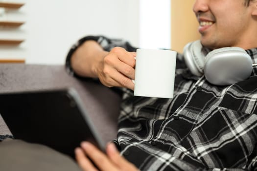 Pleased young man holding digital tablet and drinking coffee relaxing on couch at home.