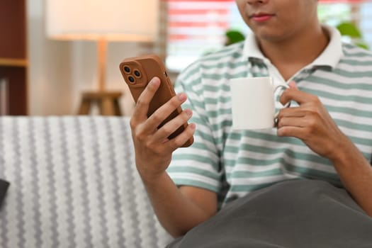 Young smiling man holding smartphone and drinking coffee relaxing on couch at home.