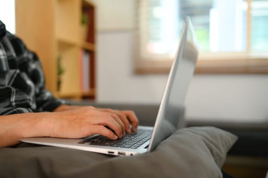 Closeup young man sitting on sofa, hands typing on laptop keyboard.