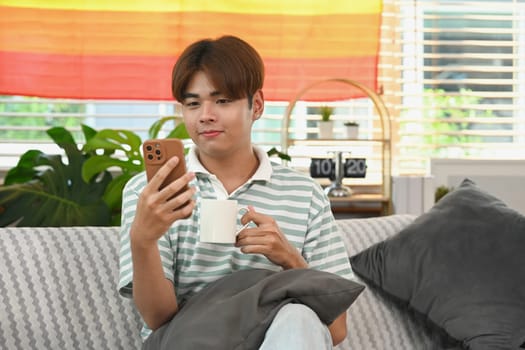 Carefree young Asian man drinking hot tea and using mobile phone on couch at home.