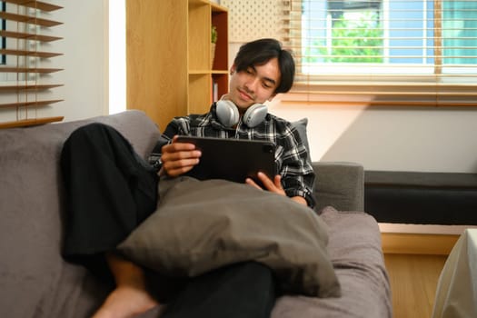 Young man relaxing on couch in living room and using digital tablet for internet surfing.