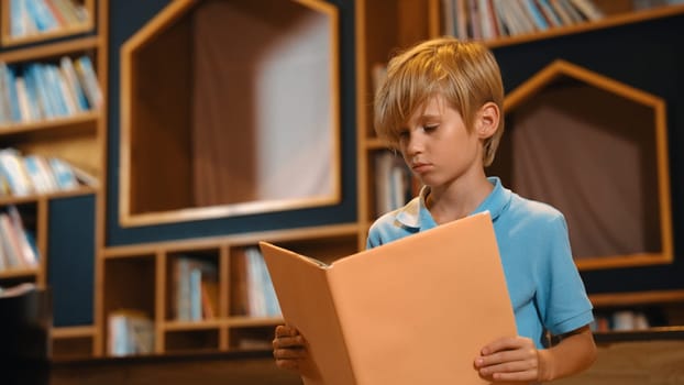 Attractive caucasian clever boy reading a book at library with stack of books placed. Smart child focus on studying, learning from novel or textbook while turning a page. Self-education. Erudition.