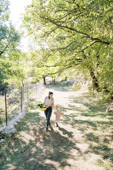 Little girl follows her mother carrying a bowl of green cabbage in her hands along the fence in a green park. High quality photo