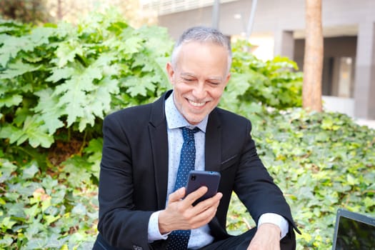 Satisfied with the results, the businessman walks down the street outside the office building, a mature boss holds a phone and coffee in his hands, writes messages and reads news online, using an app.