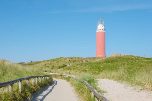 A winding path through the sandy dunes leads to a majestic lighthouse standing tall against the blue sky, guiding lost sailors home. The iconic red lighthouse of Texel Netherlands