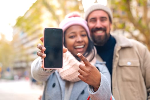 Multiracial tourist couple shows a smartphone with an empty screen for advertising or presentation, look at the camera, smiling. Gadget Concept