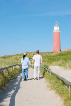 Two individuals strolling along a scenic path near a majestic lighthouse in Texel, Netherlands, surrounded by the calming sounds of the ocean in the sand dunes