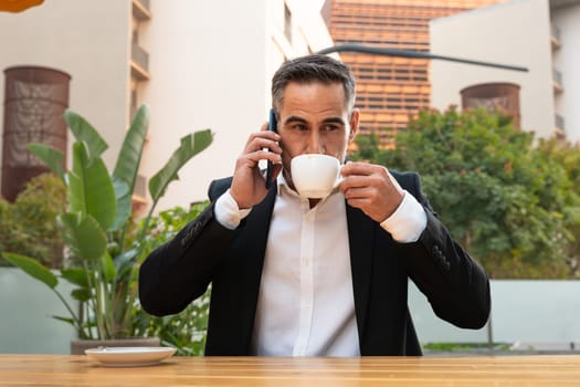 Middle aged man sitting drinking coffee happily having a work conversation with cell phone.