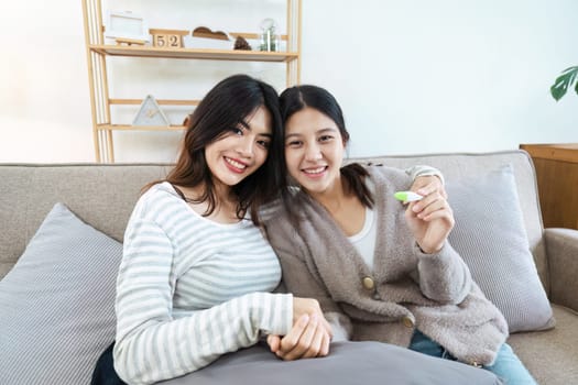 Two happy gay lesbian lgbt women couple sitting on sofa celebrating positive pregnancy test. lgbtq concept .