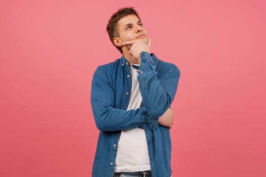 Young attractive and cheerful man of mind touching chin and deciding background isolated on pink color